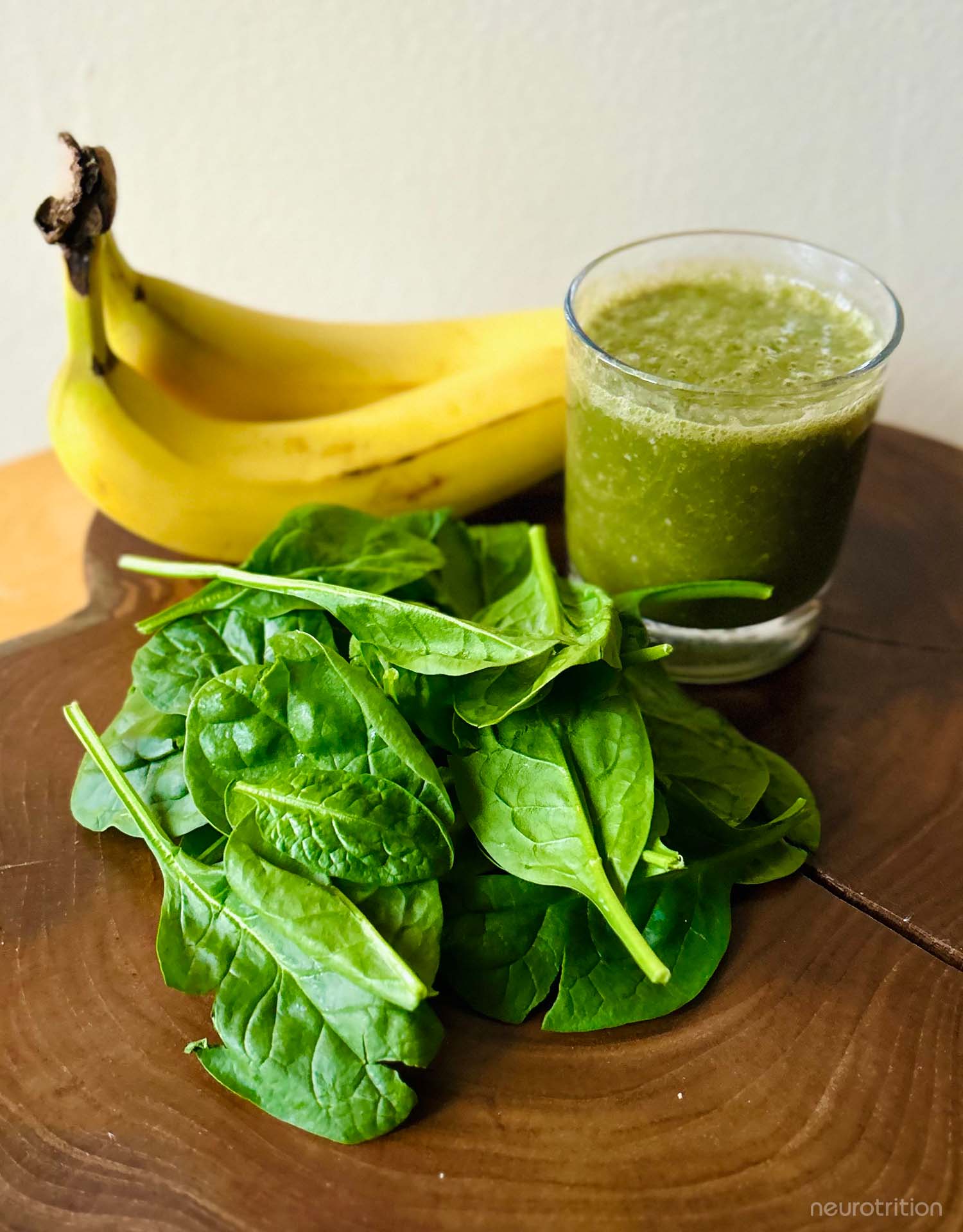 The “Green Brain” Smoothie A green smoothie with bananas behind the glass and raw spinach sprinkled with hemp seeds in front, on a wooden board.