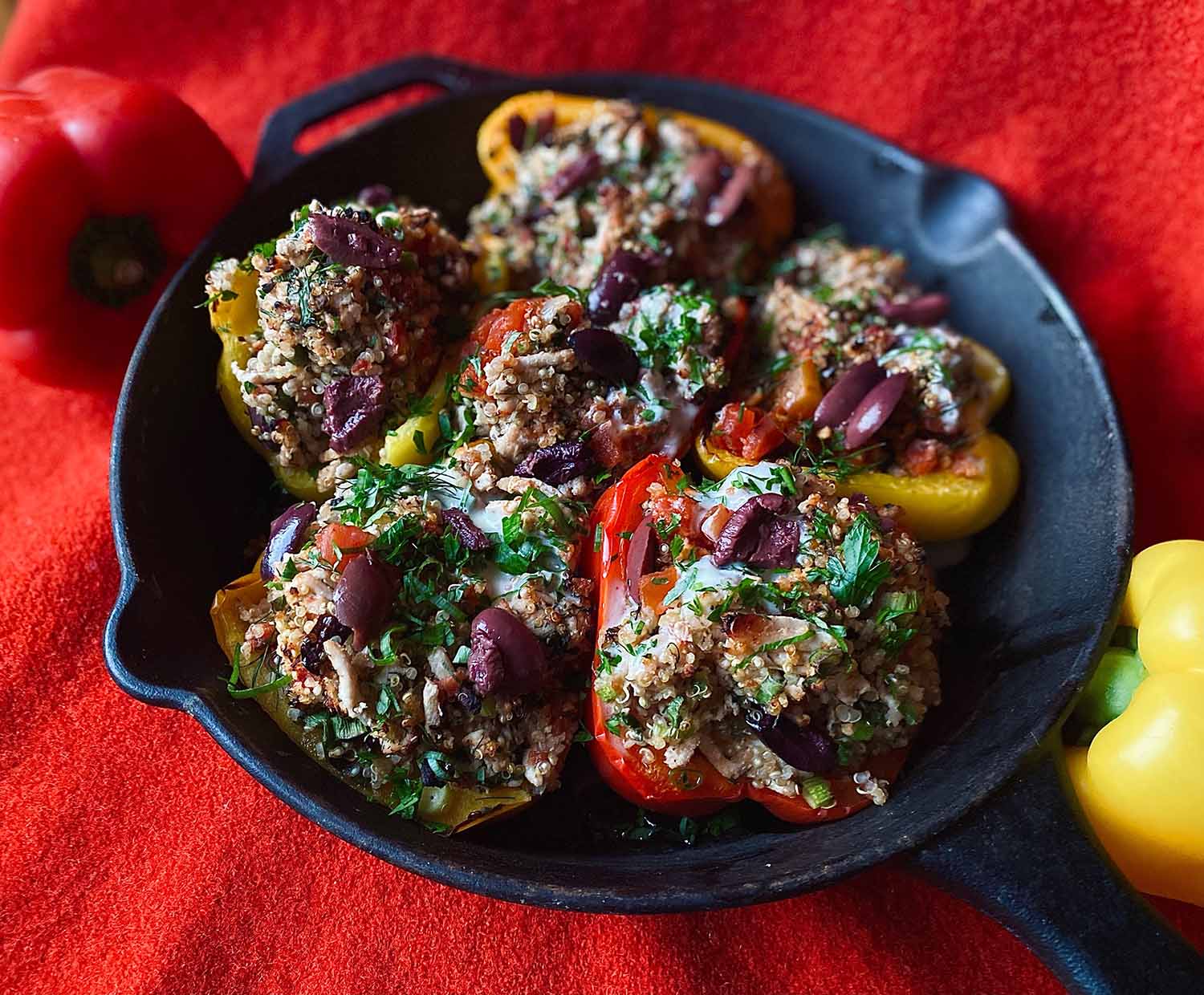 Turkey Stuffed Peppers Red and yellow bell peppers stuffed with turkey, quinoa, olives, and herbs, in a cast iron pan, placed on a red linen background.