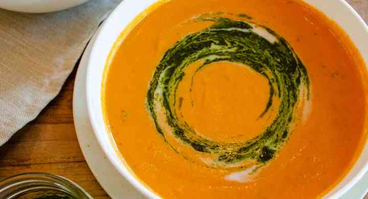 Creamy Roasted Tomato Soup Two white bowls of roasted tomato soup sit on a wooden top table, with fresh whole tomatoes and a jar of pesto beside.