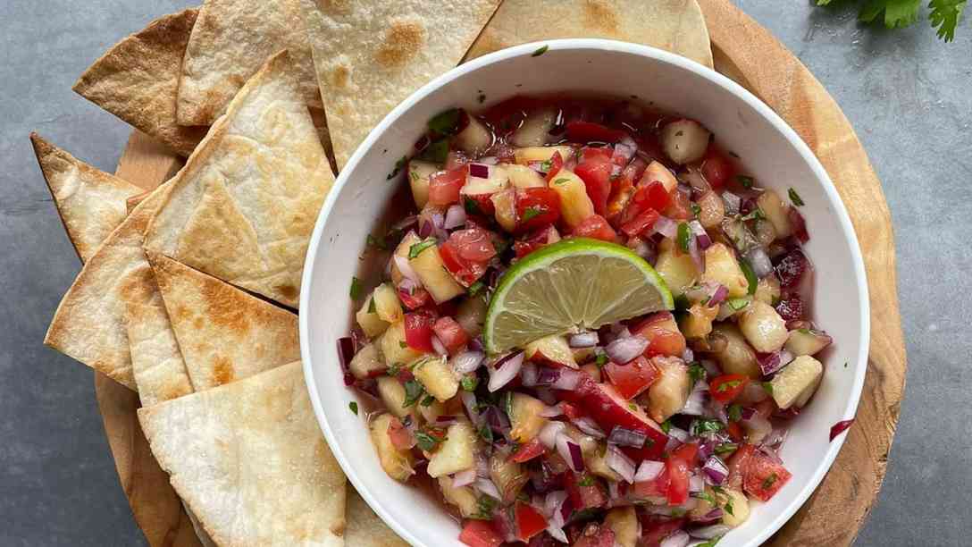 A bowl of homemade salsa is surrounded by baked pita chips on a wooden tray.